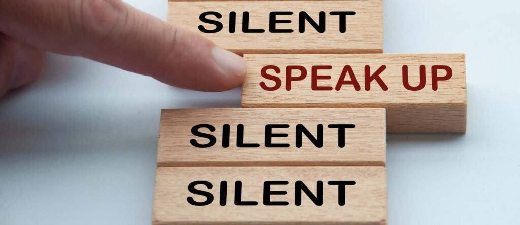 A person’s finger pushes a wooden block labeled “Speak Up” in red, moving it away from a row of blocks labeled “Silent,” symbolizing breaking the silence around workplace misconduct.