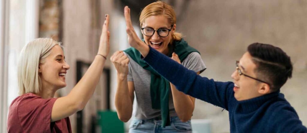 Smiling intern high-fives manager while a teammate cheers in the background — celebrating a shared workplace win.